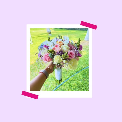 Hand holding a colorful bridal bouquet with roses, purple and white flowers against a green field background