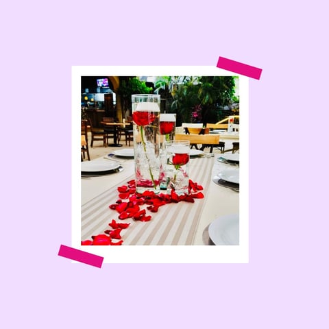 Polaroid-style photo of elegant outdoor dining table decorated with red rose petals and white floral centerpiece, held with pink tape on lavender background