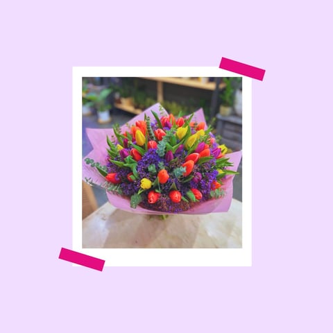 Colorful bouquet of red, yellow, and purple flowers with green foliage wrapped in pink paper, pinned to lavender background