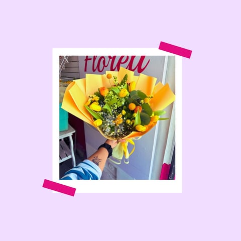 Person holding a bouquet of yellow flowers wrapped in yellow paper with green foliage inside, pinned to a lavender background