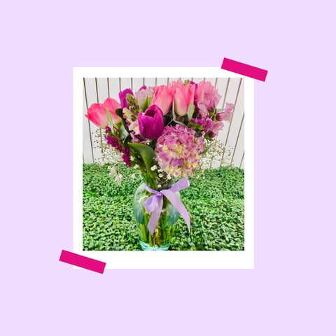 Polaroid-style photo of pink and magenta flowers in a vase with lavender bow on light purple background
