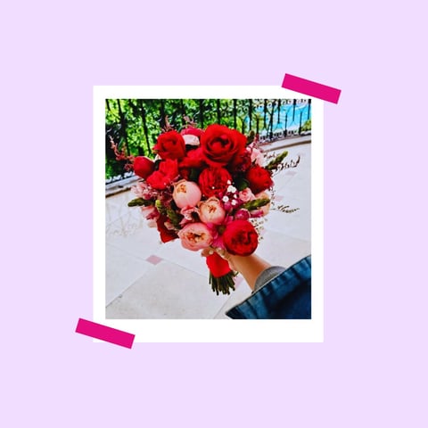 Polaroid-style photo of a vibrant red and coral rose bouquet held in someones hand against a purple background with pink tape