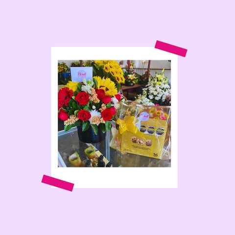 Polaroid-style photo of vibrant flower arrangements with red roses and sunflowers, along with packaged items, held by pink tape on lavender background